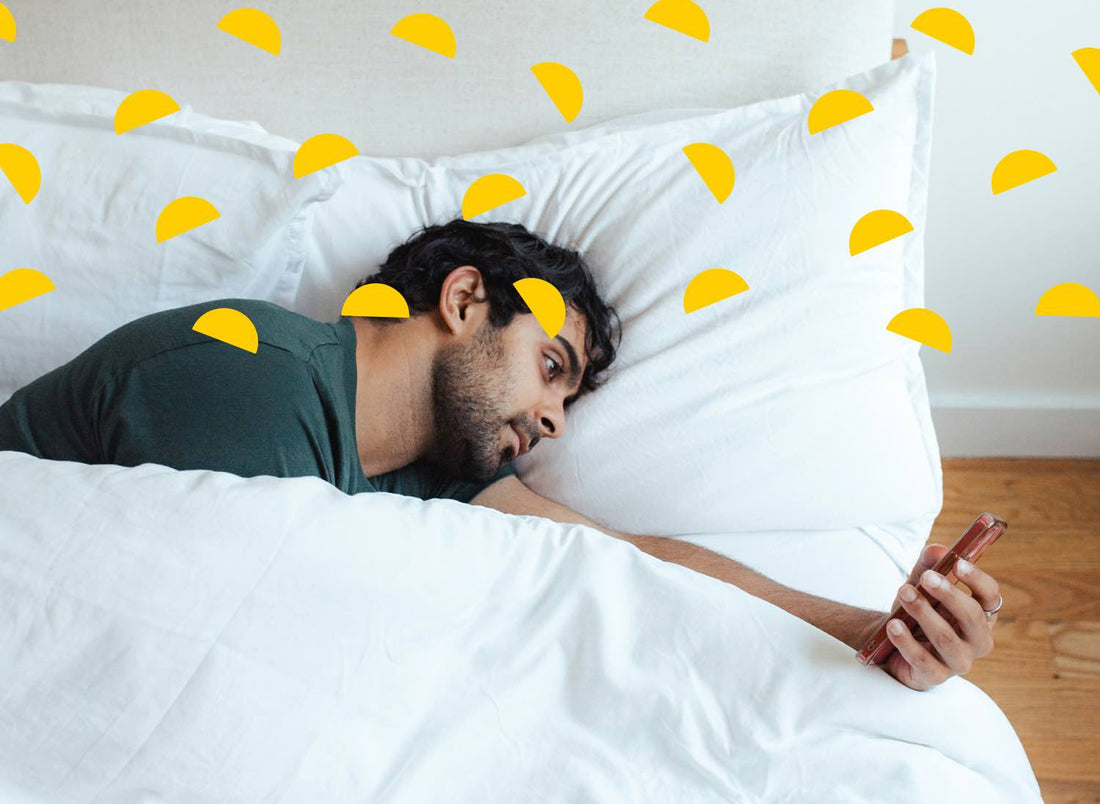 A man lying in bed on his side, looking at his phone with a bored expression on his face.