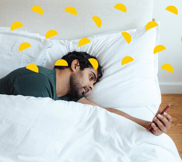 A man lying in bed on his side, looking at his phone with a bored expression on his face.