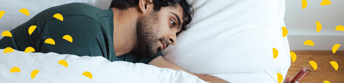 A man lying in bed on his side, looking at his phone with a bored expression on his face.