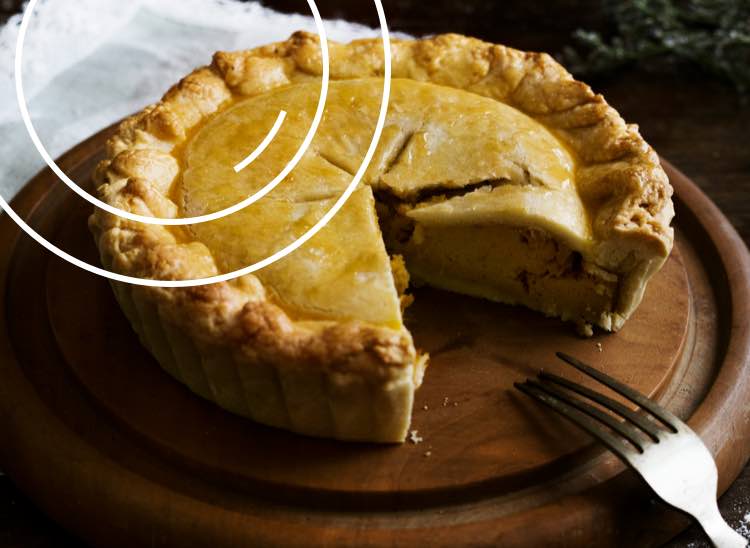 A tasty savoury pie sitting on a round wooden board with a slice missing and a fork where it once was.